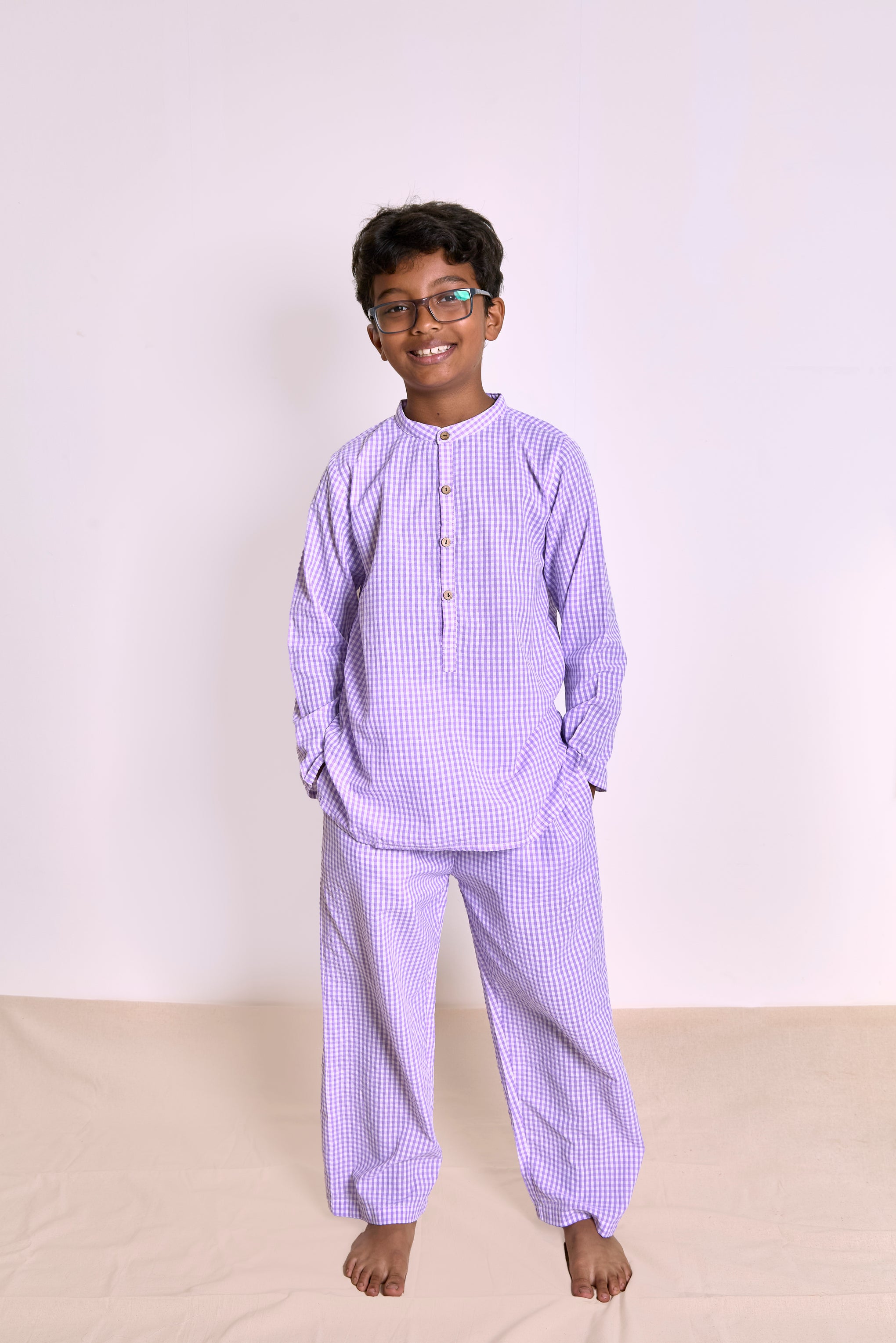 A young boy wearing a light purple organic cotton seersucker two-piece pajama set, consisting of a long-sleeve top with a button placket and matching pants, standing against a plain light background.